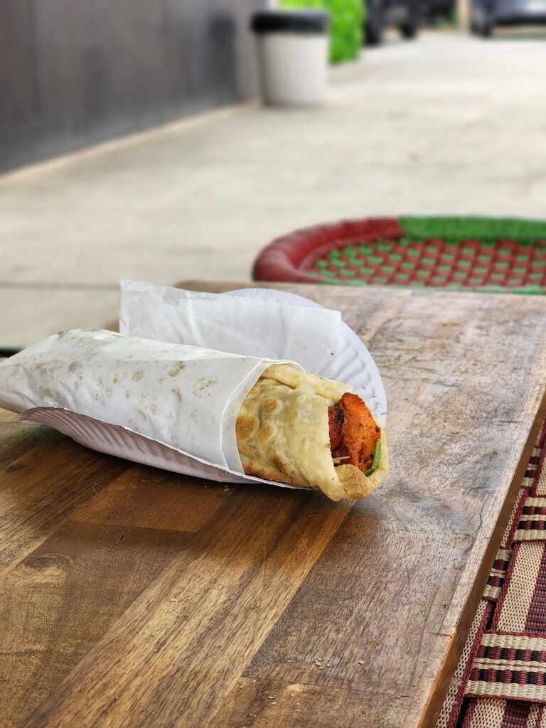 Chicken tikka roll wrapped in paper on a wooden table beside a woven manjaa